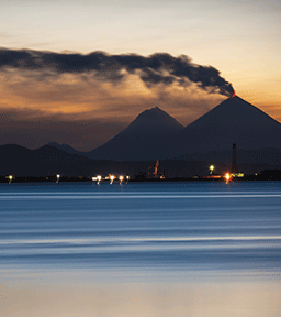 Volcano erupting at sunset over a calm sea, with smoke and glowing lava visible against the twilight sky.