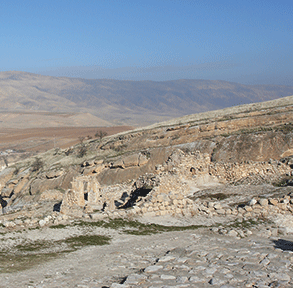 Ancient ruins on a rocky hillside with a backdrop of arid mountains under a clear blue sky.