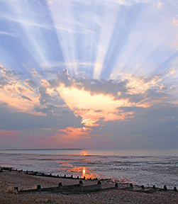Sunrise over a calm beach with dramatic sun rays piercing through clouds, reflecting on the ocean.