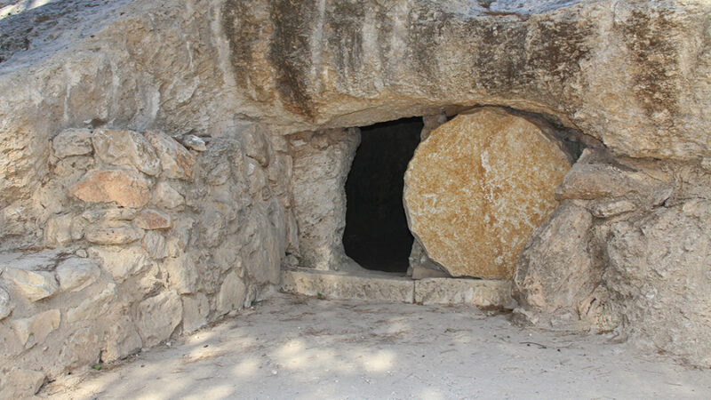 Ancient stone tomb entrance with rolled stone door surrounded by rocky terrain.