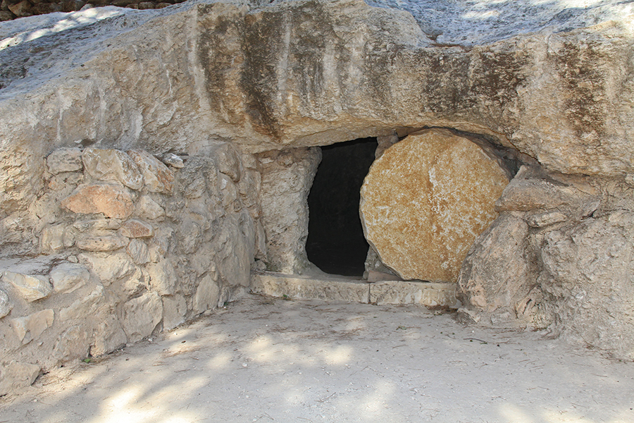 Ancient stone tomb entrance with rolled stone door surrounded by rocky terrain.