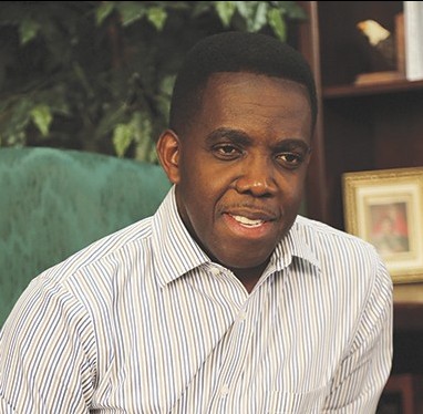 Man smiling indoors, wearing striped shirt, sitting in a room with framed photo and plants in the background.