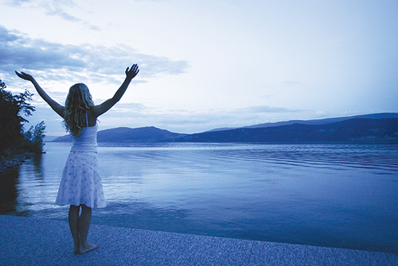 Woman embracing serene lakeside view at sunset, arms raised in peaceful gesture, surrounded by calm water and mountains.