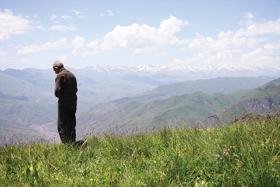 Man observing scenic mountain landscape with green valley and snowy peaks under a blue sky with clouds.