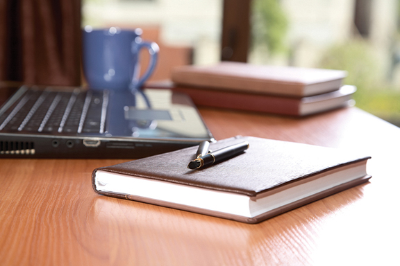 Notebook and pen on a wooden desk beside a laptop and coffee cup, perfect for productivity and workspace inspiration.