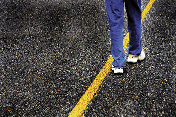 Person walking on a road's yellow dividing line, wearing blue pants and sneakers, symbolizing journey and balance.