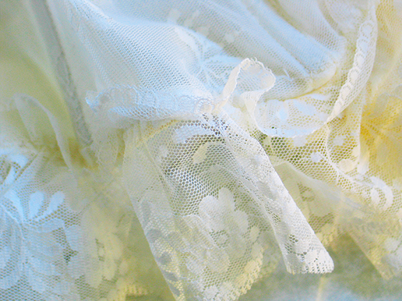 Delicate white lace fabric with floral patterns, draped elegantly in soft folds, highlighting intricate embroidery.