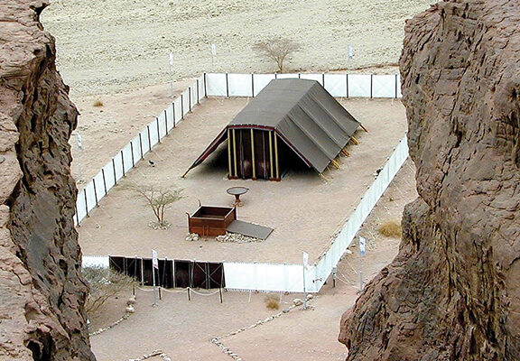 Replica of ancient Tabernacle in desert landscape, surrounded by fencing, viewed from between rocky cliffs.