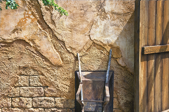 Old wooden wheelbarrow against a rustic, textured wall with cracked paint and a wooden door in the sunlight.
