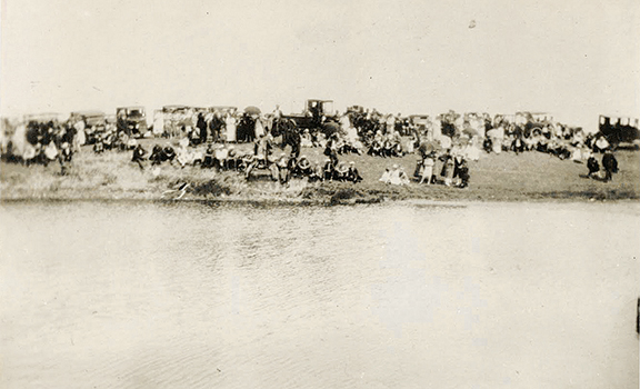 Vintage gathering by a lakeside, with numerous people and early 1900s cars in the background.