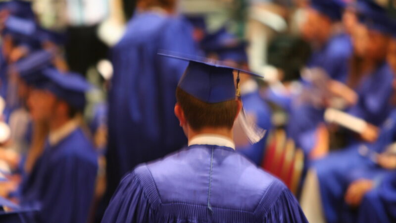 Graduate in blue cap and gown at commencement ceremony, facing stage.