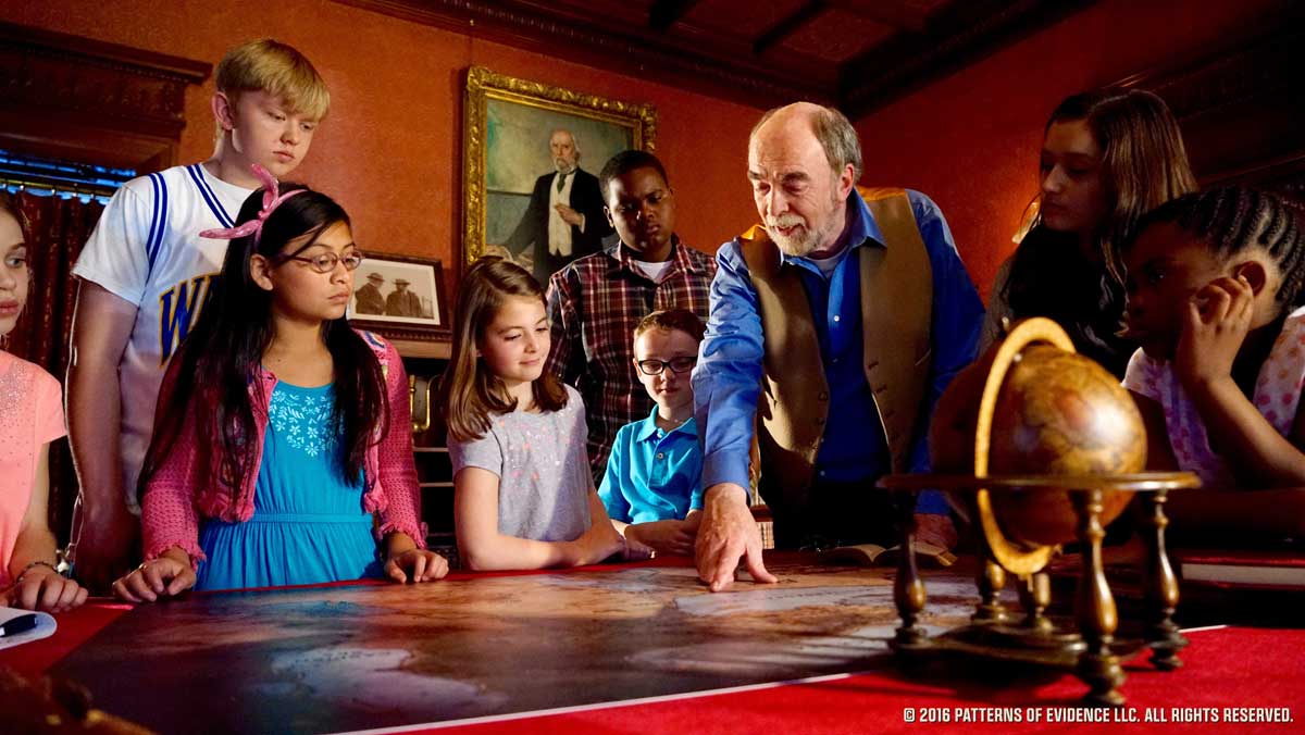 Group of children and man examining a map on a table in a dimly lit room with a globe nearby.