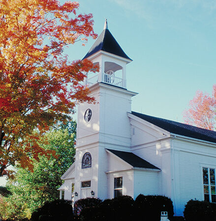 White church with steeple amidst autumn trees and clear sky, showcasing serene fall scenery.