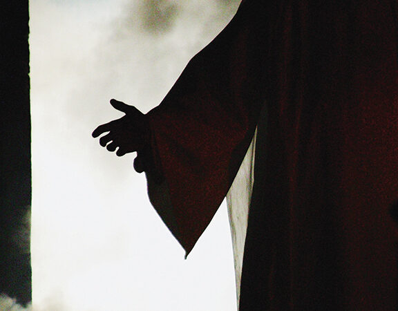 Silhouetted outstretched hand against a bright, cloudy background, evoking a sense of welcome or invitation.