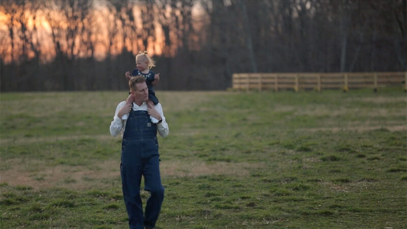 Father carrying child on his shoulders in a grassy field at sunset, wearing overalls, with trees in the background.