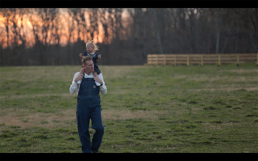 Father carrying child on his shoulders in a grassy field at sunset, wearing overalls, with trees in the background.