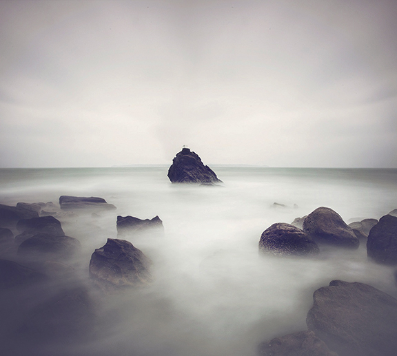 Mysterious seascape with misty waves, isolated rock formations, and overcast sky creating a serene, ethereal atmosphere.