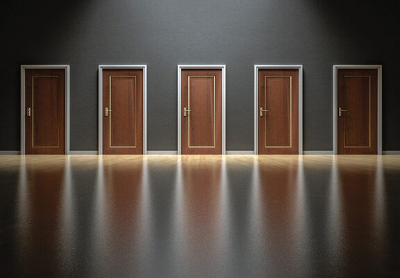 Five closed wooden doors in a dimly lit room with reflections on the shiny floor, symbolizing choices and decisions.