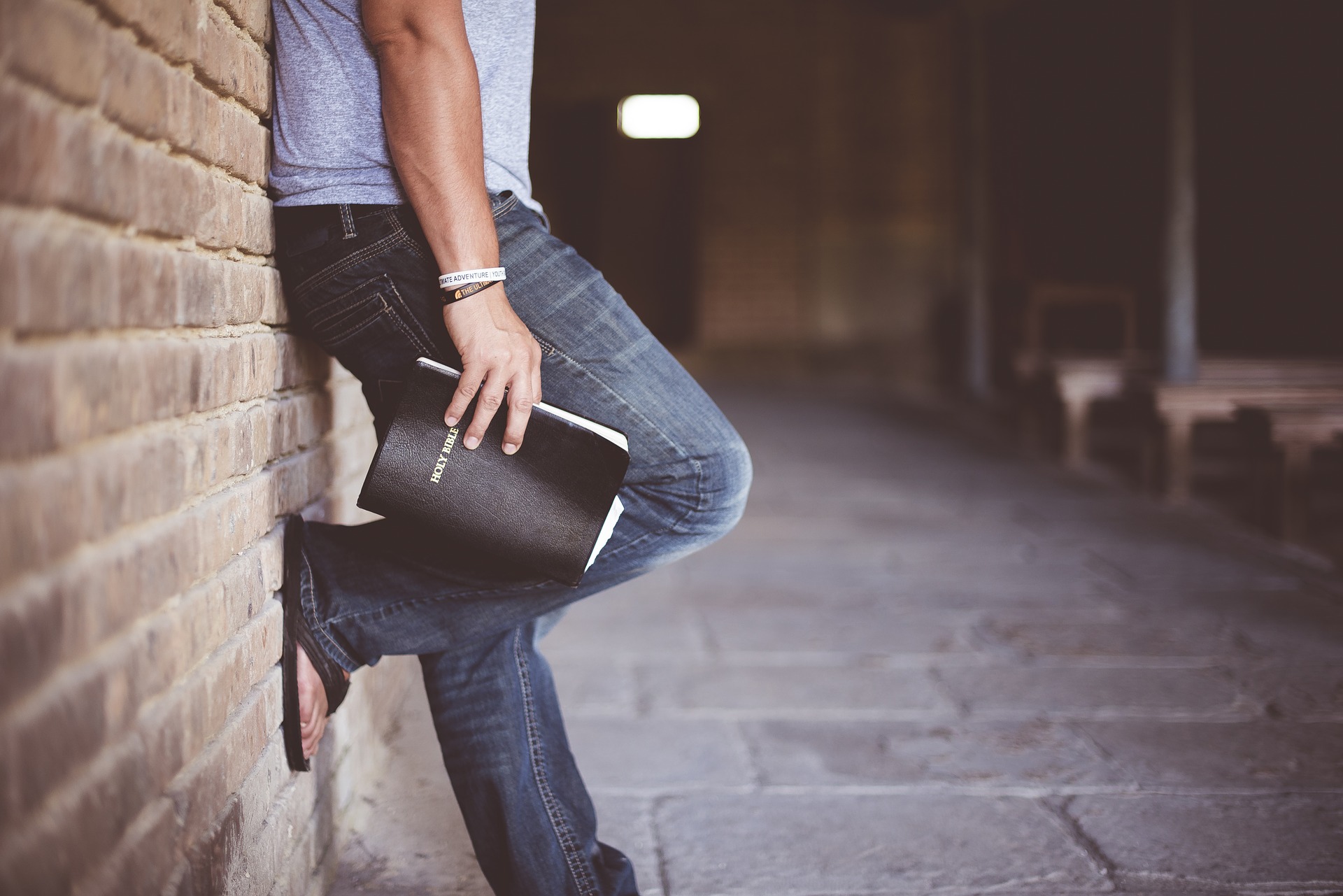 Person leaning on wall holding a Bible, wearing jeans and flip-flops, suggesting reflection or spirituality.