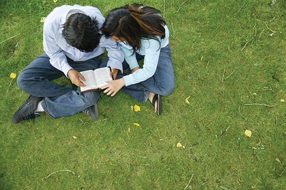 Two people sitting on grass reading a book together, captured from above.