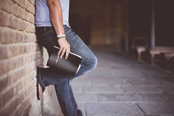 Person leaning against a brick wall holding a book, wearing casual jeans and a gray shirt, on a rustic stone path.