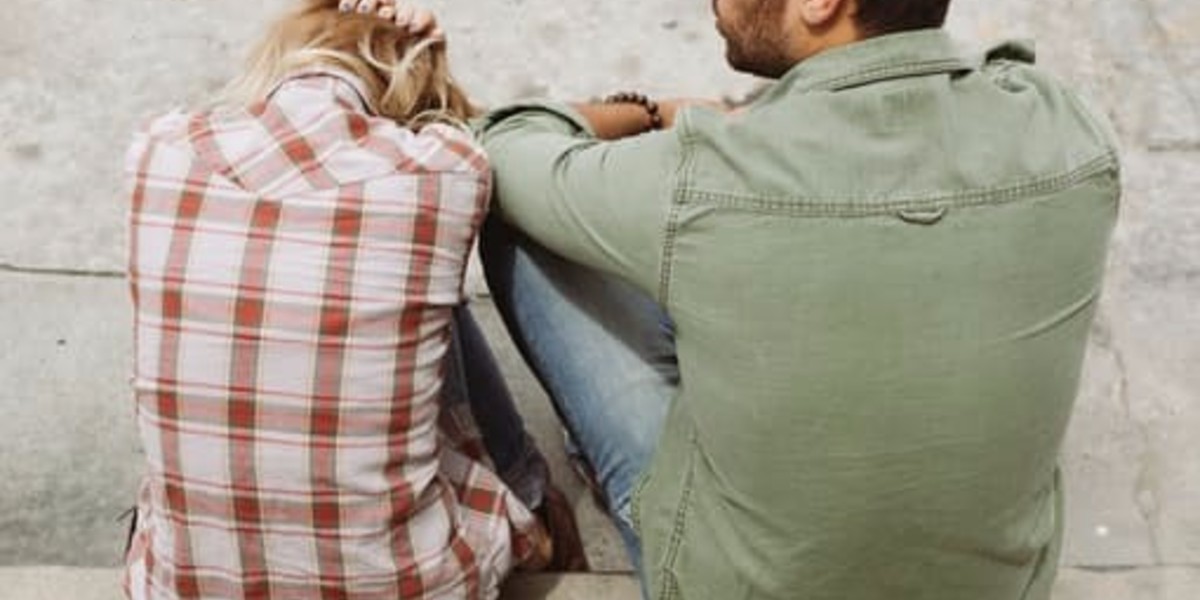 Couple sitting on a sidewalk, seen from behind. Woman wears plaid shirt; man in green jacket, both in conversation.