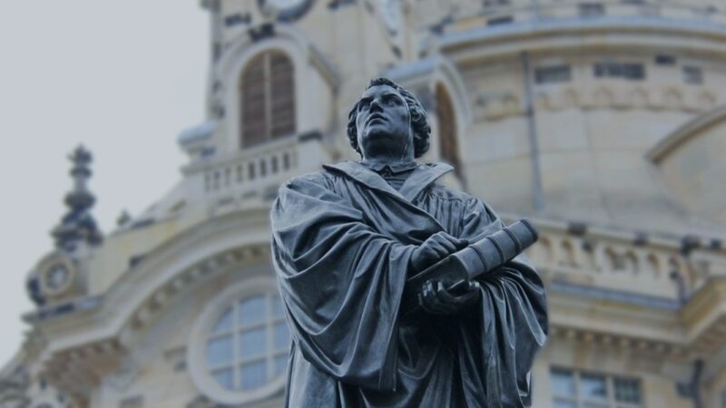 Statue of a historical figure holding a book in front of a detailed architectural building.