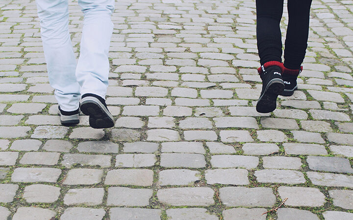 Two people walking on cobblestone street, wearing casual pants and shoes, captured from behind.