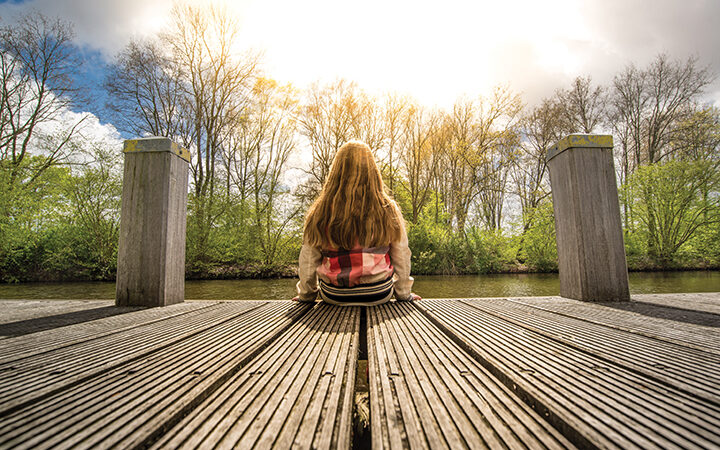 Person sitting on a wooden pier, gazing at a serene river under a bright sky, surrounded by trees in spring.