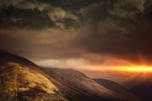 Dramatic mountain landscape at sunset with dark clouds and a vibrant orange sun peeking through.