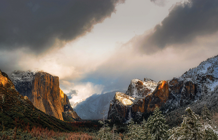 Majestic view of Yosemite Valley's El Capitan and Half Dome enveloped in dramatic clouds and soft sunlight.