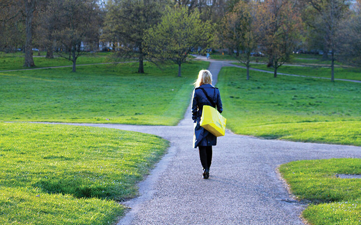 Woman walking in a park on a sunny day, carrying a yellow bag and wearing a blue coat.