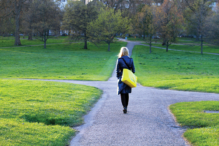 Woman walking in a park on a sunny day, carrying a yellow bag and wearing a blue coat.