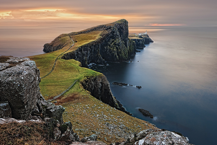 Stunning coastal cliff with winding path and distant lighthouse under a dramatic sky at sunset.