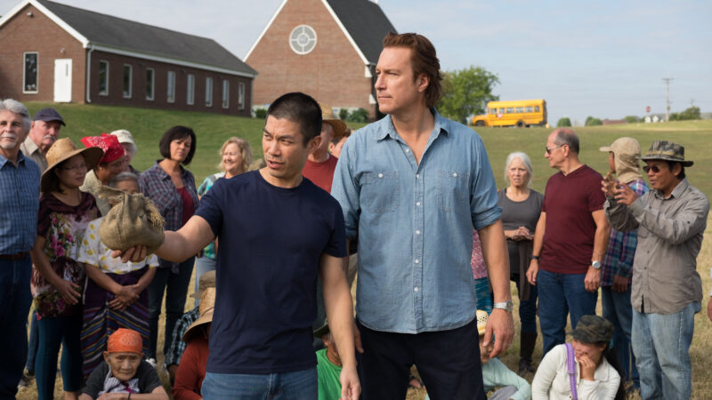 Community gathering in front of a church, man holding a burlap sack, diverse group listening attentively, bus in background.