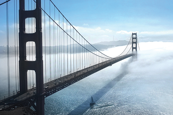 Golden Gate Bridge enveloped in fog, with a sailboat below on a sunny day.