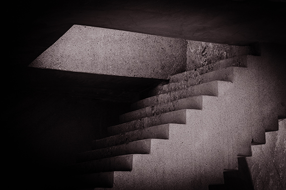 Dimly lit concrete staircase with sharp angles leading to a rectangular skylight, casting dramatic shadows.
