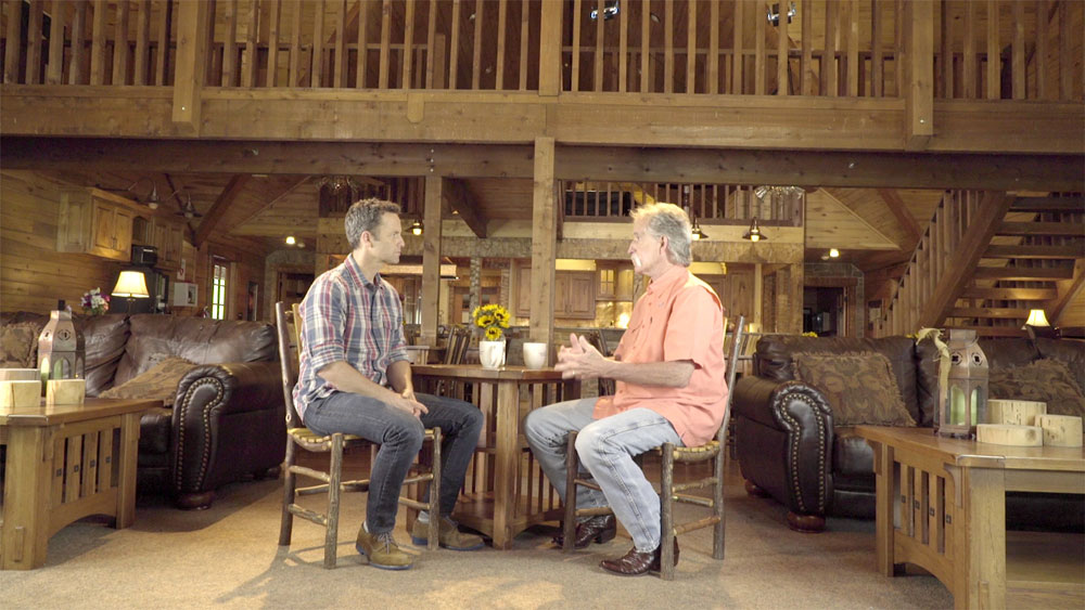 Two men having a conversation in a cozy wooden cabin living room with rustic furniture and a warm atmosphere.