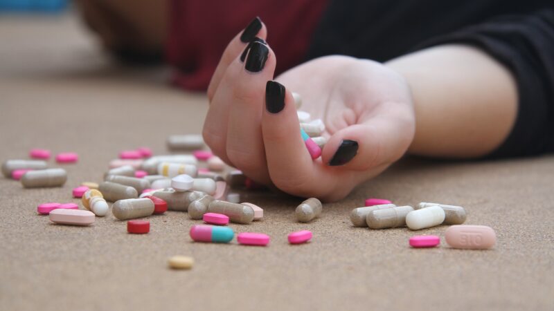 Hand with black nails amidst scattered colorful pills on surface.