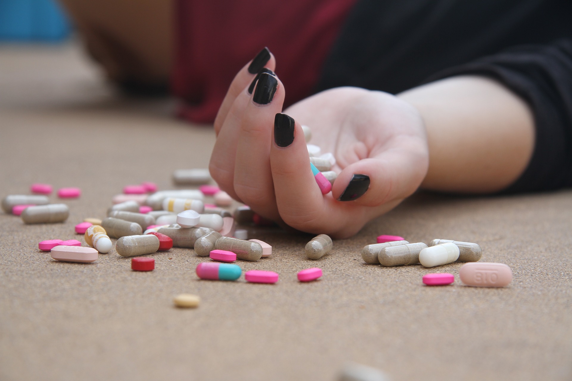 Hand with black nails amidst scattered colorful pills on surface.