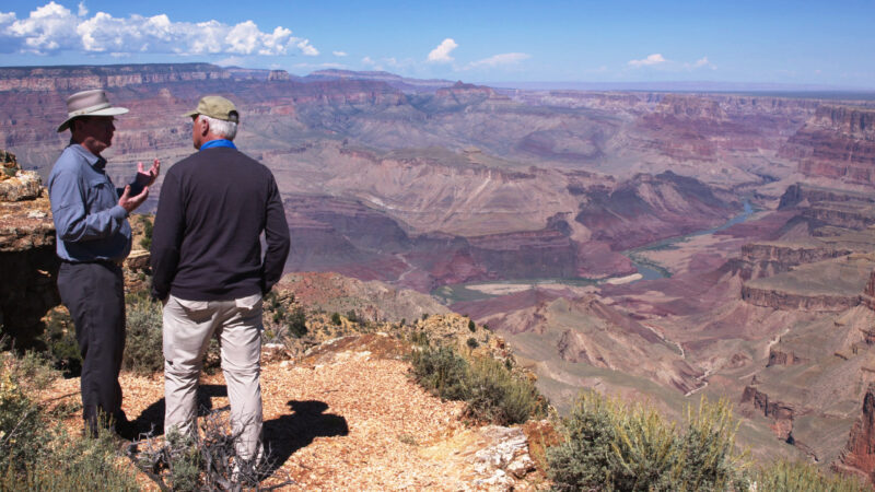 Two men talking at Grand Canyon overlook with expansive rocky landscape and river below.