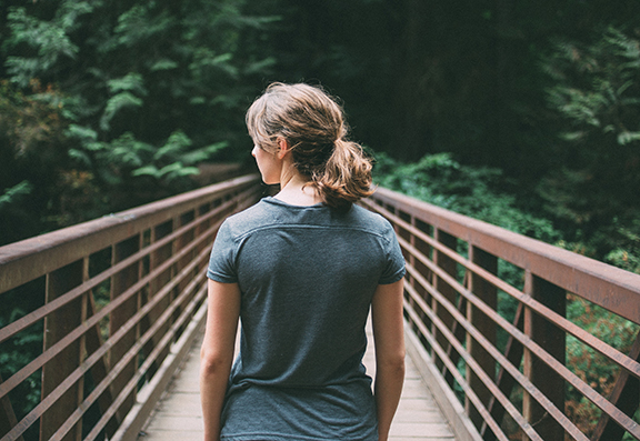 Woman walking on a wooden bridge in a lush forest, viewed from behind, wearing a gray shirt and ponytail.