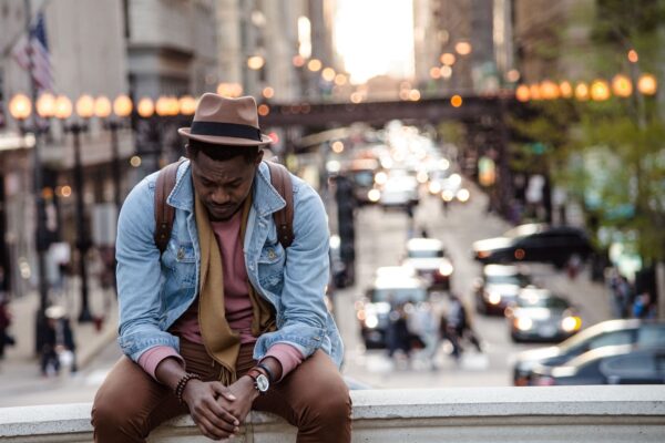 Man sitting pensively on city street ledge, traffic blurred in background, wearing denim jacket and fedora hat.