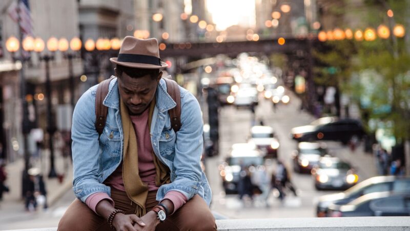 Man sitting pensively on city street ledge, traffic blurred in background, wearing denim jacket and fedora hat.