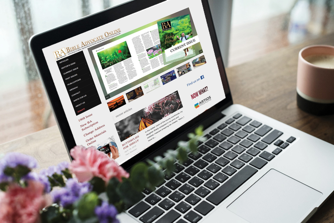 Laptop displaying Bible Advocate Online magazine, surrounded by flowers, with a coffee mug on a wooden table.