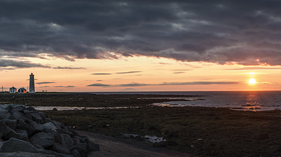 Lighthouse on rocky shore at sunset with dramatic clouds and ocean view.