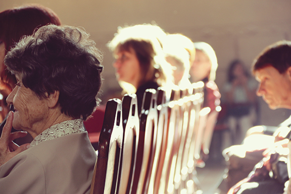 Elderly woman and others seated in a row of wooden chairs, sunlight illuminating faces, creating a thoughtful atmosphere.