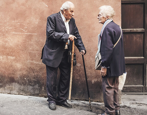 Elderly couple conversing in the street, both using canes; man in suit, woman in scarf and coat.