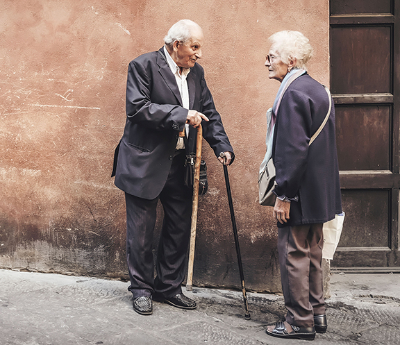 Elderly couple conversing in the street, both using canes; man in suit, woman in scarf and coat.