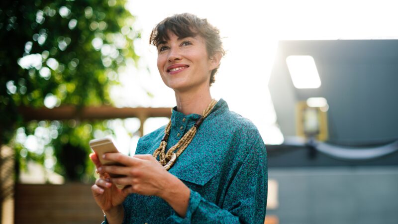 Smiling woman in green shirt uses smartphone outdoors, bathed in sunlight.